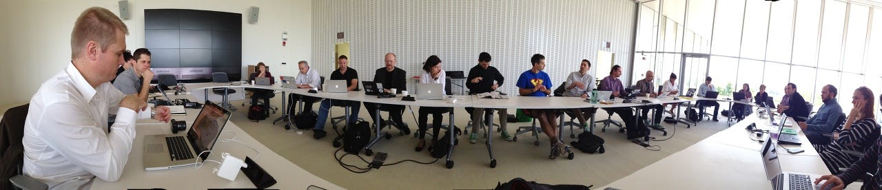 A wide panorama of about twenty people seated at long white tables with laptops in a bright, glass-walled room.