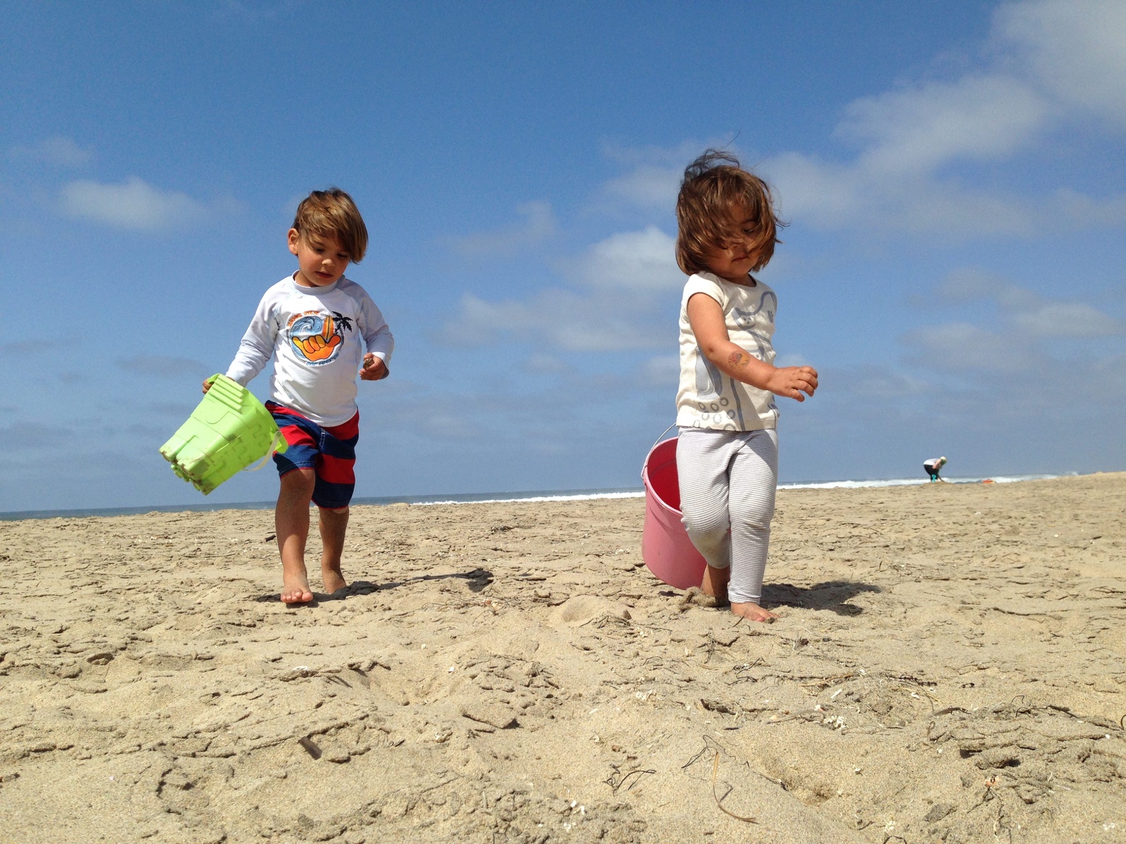 Two young children walking across a sandy beach carrying plastic buckets, the ocean and a blue sky behind them.